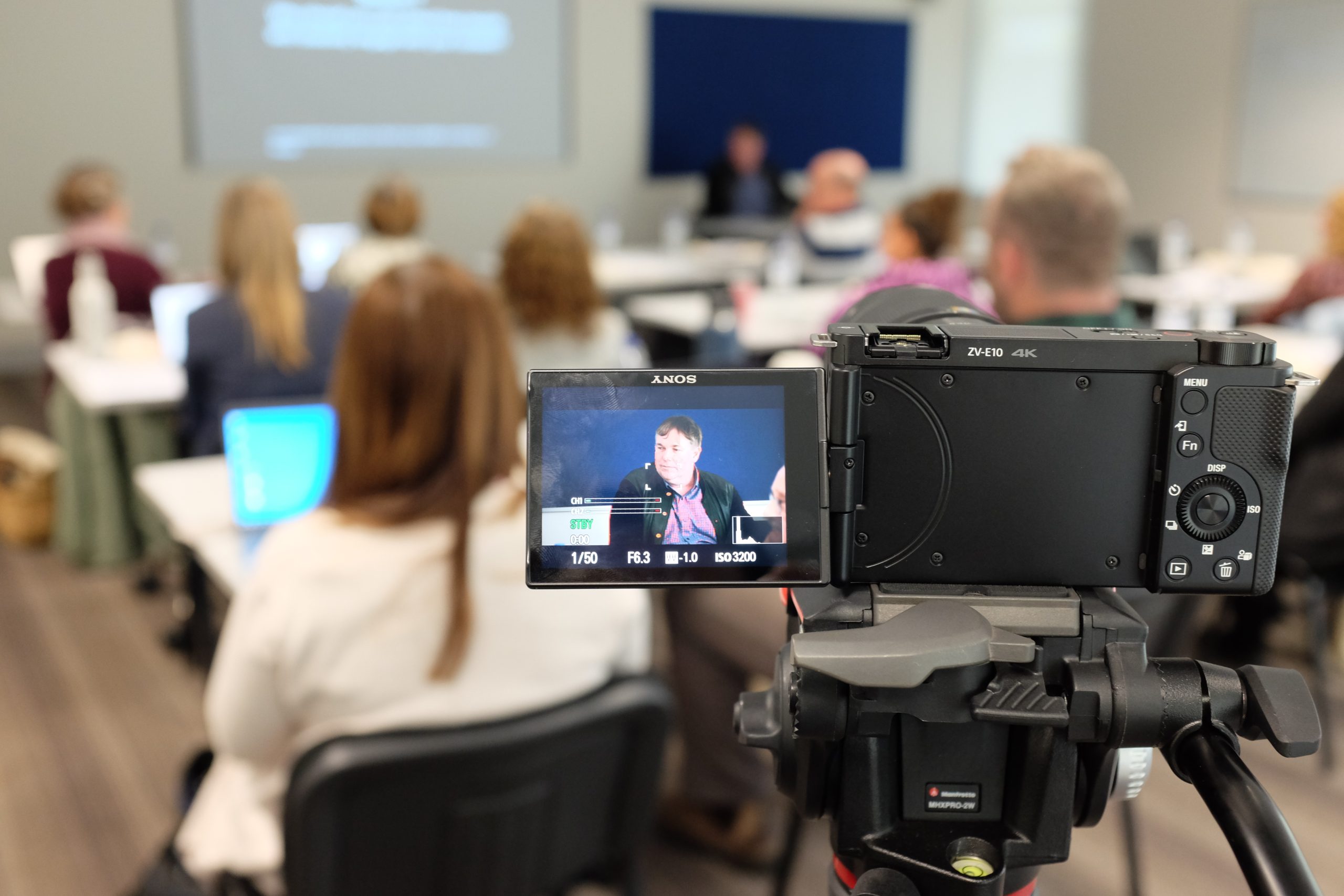 Image is of a film camera. The picture on the camera is Dr. Scott Hollier presenting. Behind the camera is the group of attendees listening to the presentation.