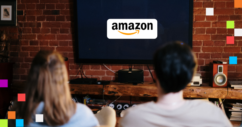 The back of two people, a man and a woman, as they watch a television screen. The screen has the Amazon logo on it.