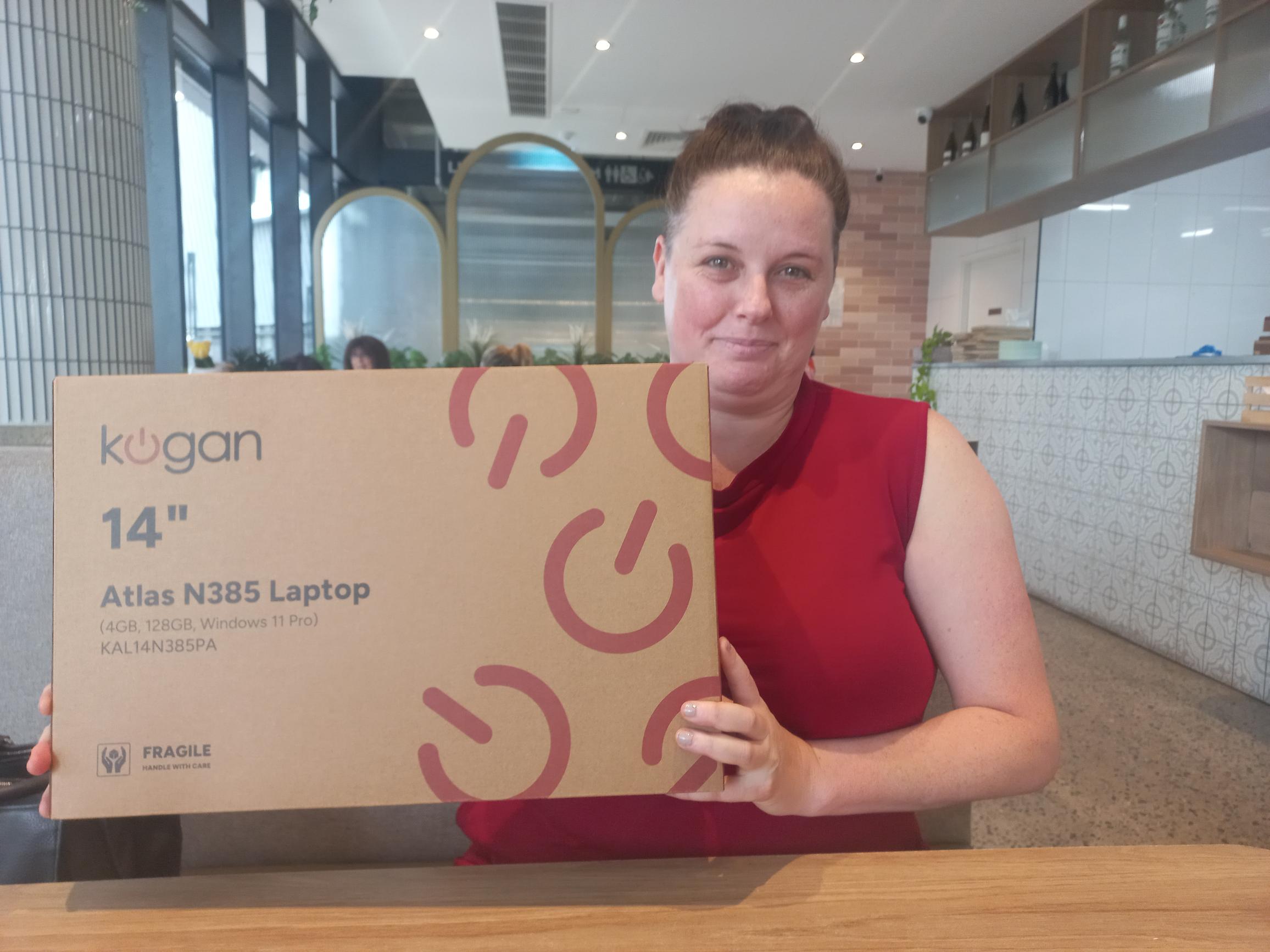 An image of a smiling woman, Emma, sitting in the booth of a cafe. She is holding a box with a laptop inside.